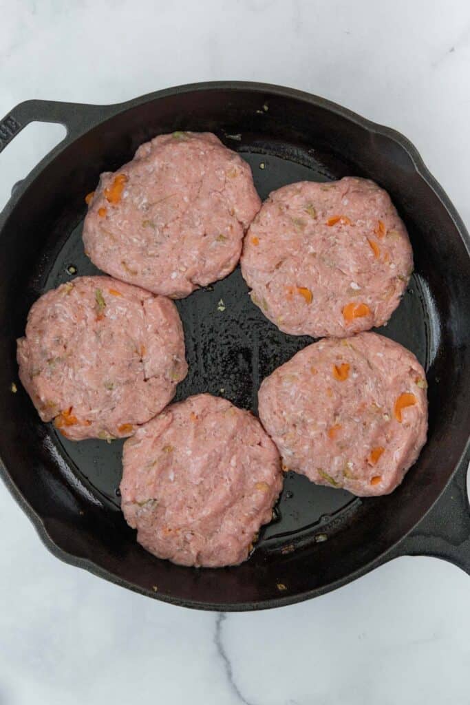 Five raw, seasoned turkey burger patties with visible diced vegetables are placed in a black cast iron skillet on a white marble surface.