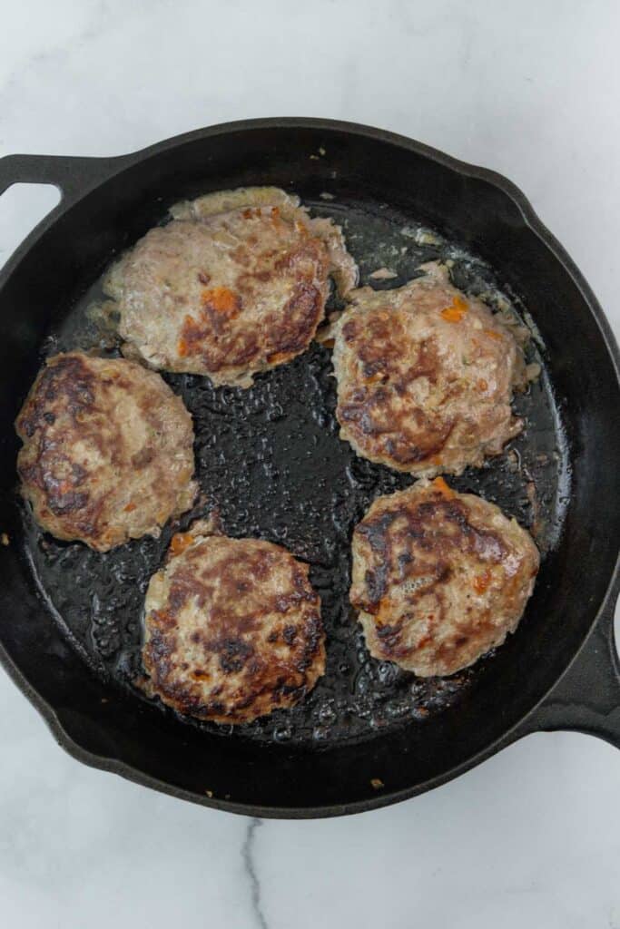 Five turkey burgers cooking in a black cast-iron skillet on a marble surface.