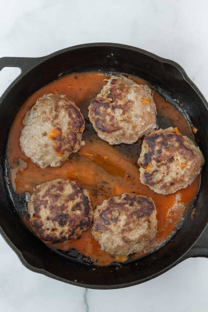 Four cooked turkey burger patties in a black cast iron skillet with buffalo sauce, viewed from above on a white marble background.