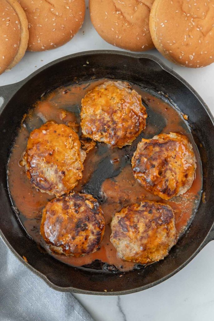 Four cooked turkey burger patties smothered in buffalo sauce in a black cast iron skillet, with sesame seed buns nearby on a white surface.