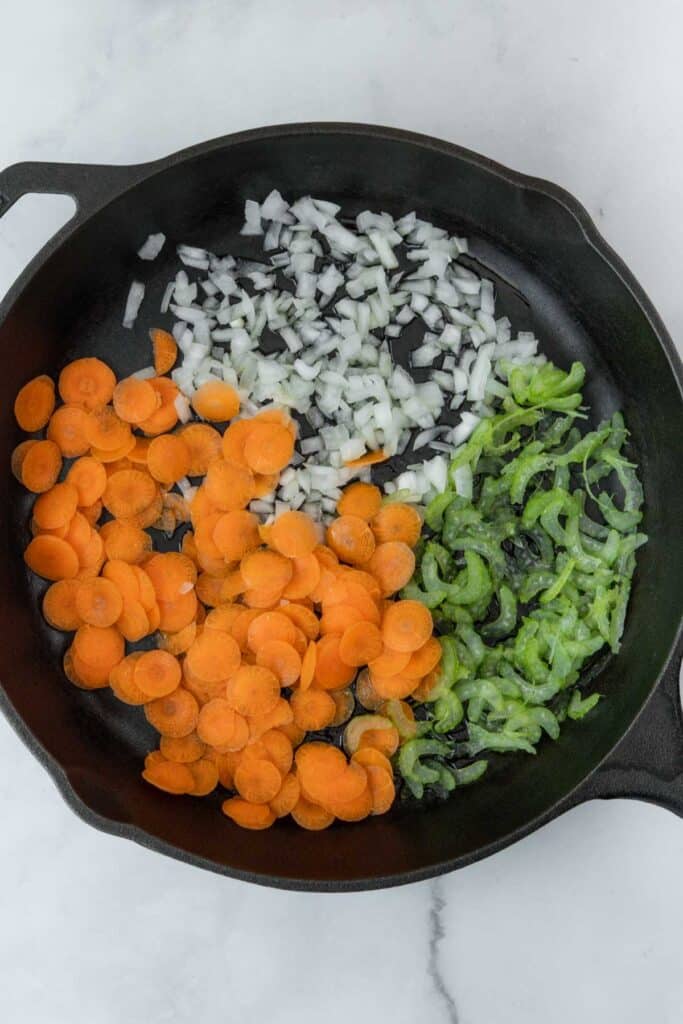 A cast iron skillet on a marble surface containing neatly arranged sliced vegetables: diced carrots, onions, and celery.