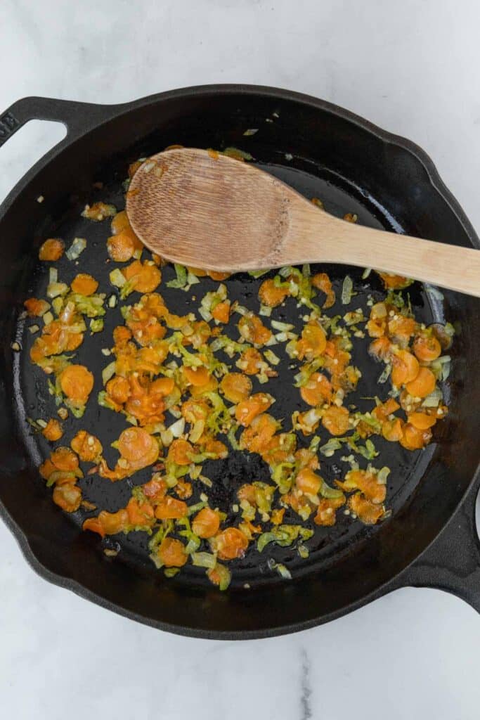 Sautéed chopped vegetables including carrots and celery in a black cast iron skillet with a wooden spoon.