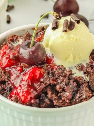 Cherry dump cake in a white bowl topped with ice cream and chocolate curls garnished with a cherry.