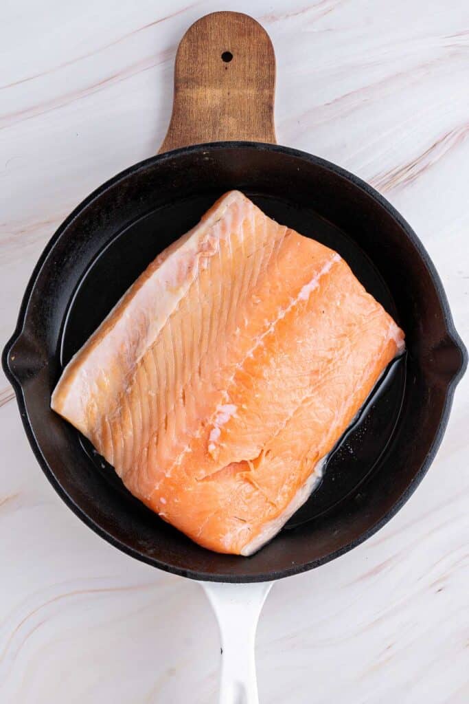 A large raw salmon fillet with visible skin is placed in a cast iron skillet.