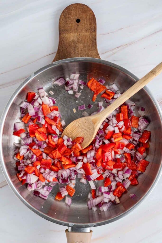 A stainless steel skillet containing chopped red onions and red bell peppers, mixed with a wooden spoon.