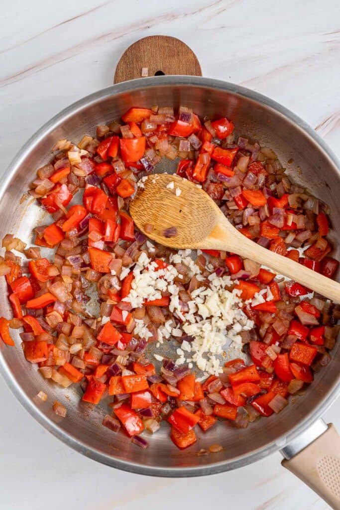 A stainless steel skillet with sautéed chopped onions and red bell peppers, a wooden spoon, and minced garlic being added.