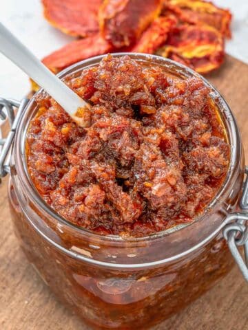 A close-up of a glass jar filled tomato jam placed on a wooden board with a spoon inserted.