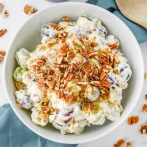 A white bowl filled with creamy grape salad topped with chopped pecans and brown sugar. Red and green grapes are visible through the creamy dressing. The bowl is placed on a light blue cloth with scattered pecans around it.