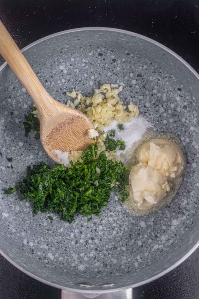 A pot containing minced garlic, chopped parsley, salt, and a dollop of butter, being stirred with a wooden spoon.