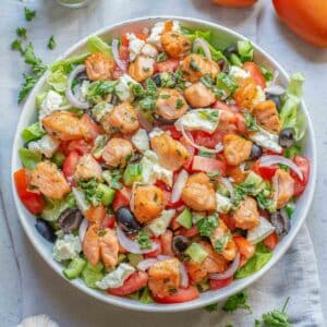 A large white bowl of greek salad with salmon with the dressing drizzled overtop.