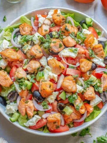 A large white bowl of greek salad with salmon with the dressing drizzled overtop.