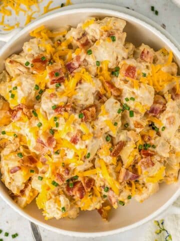 A white bowl of ranch potato salad topped with shredded cheese, crispy bacon bits, and chopped chives.