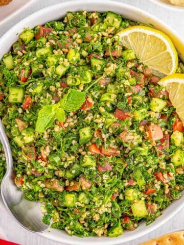 A white bowl of tabouli salad garnished with lemon slices and mint leaves on a white table and a spoon rests inside the bowl.