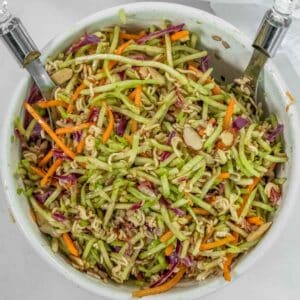 A bowl of an easy broccoli slaw recipe with sliced almonds, shredded carrots, cabbage, and green vegetables. Two silver serving utensils are placed in the bowl.