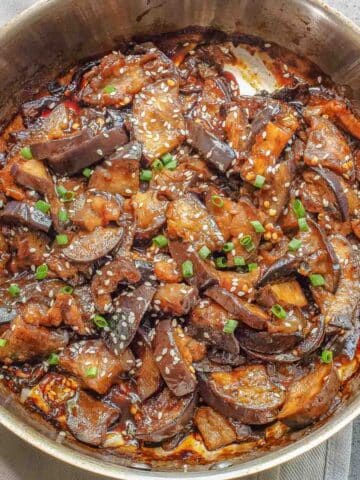A skillet filled with cooked chinese eggplant slices in a savory sauce, garnished with sesame seeds and chopped green onions.