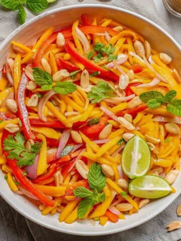 Mango salad with sliced red peppers, fresh mint, lime wedges, and roasted peanuts in a large white bowl.