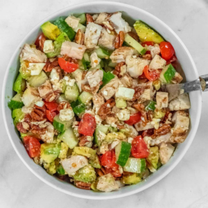 A bowl filled with a salad consisting of diced chicken, cucumbers, tomatoes, avocado, almonds, and cheese crumbles.