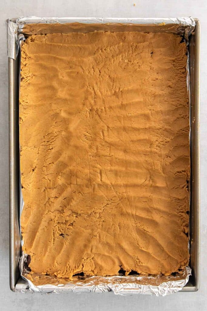 A rectangular baking pan lined with foil contains an unbaked layer of peanut butter.