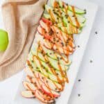 A rectangular white platter holds alternating slices of red and green apples topped with drizzle of caramel and sprinkled with chocolate chips.