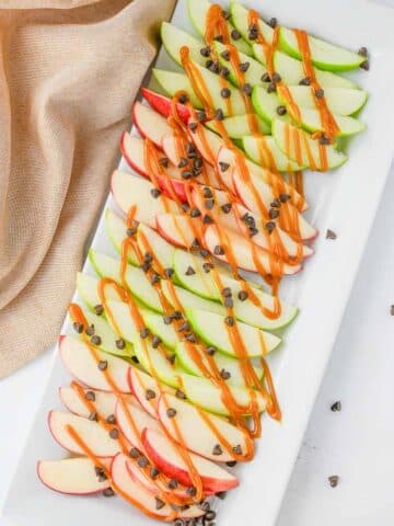 A rectangular white platter holds alternating slices of red and green apples topped with drizzle of caramel and sprinkled with chocolate chips.