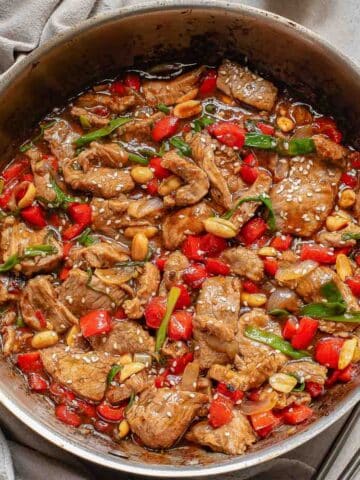 A skillet filled with cooked beef strips, red bell peppers, green onions, and peanuts in a rich brown sauce.