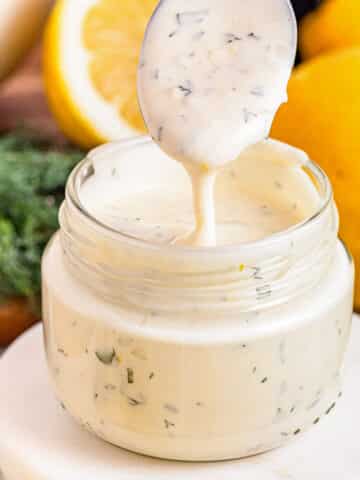 A glass jar filled with creamy lemon aioli, with a spoon being lifted out, and fresh lemons in the background.