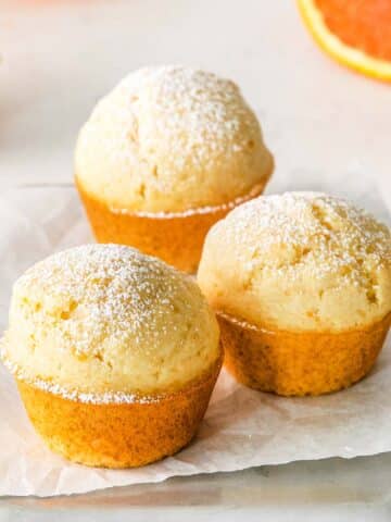 Three orange muffins, dusted with sugar, are placed on parchment paper on top of a tray.