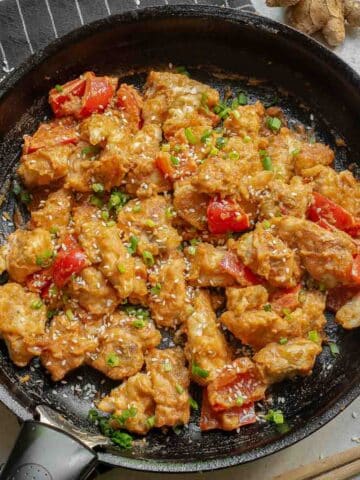 A black skillet containing a peanut butter chicken, red bell peppers, and green onions, garnished with sesame seeds.