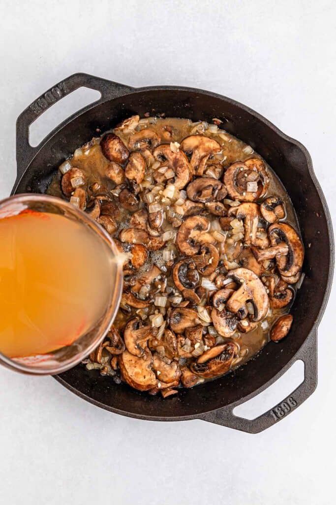 A cast iron skillet with sautéed mushrooms and onions with chicken broth being poured into the skillet.