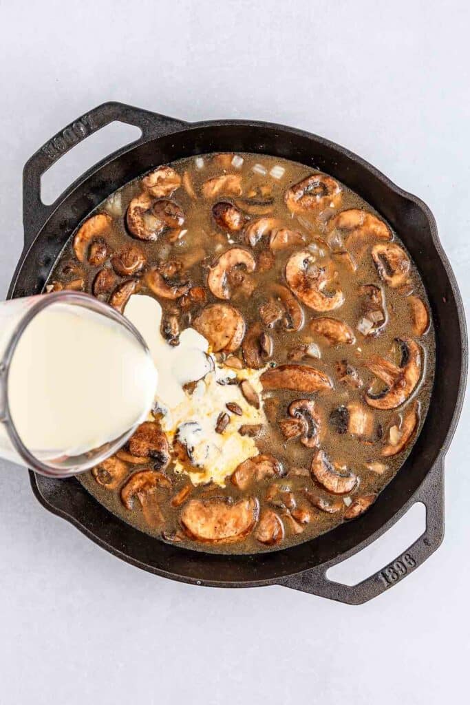 heavy cream is being poured into a black cast iron skillet filled with cooked mushrooms and chicken broth.