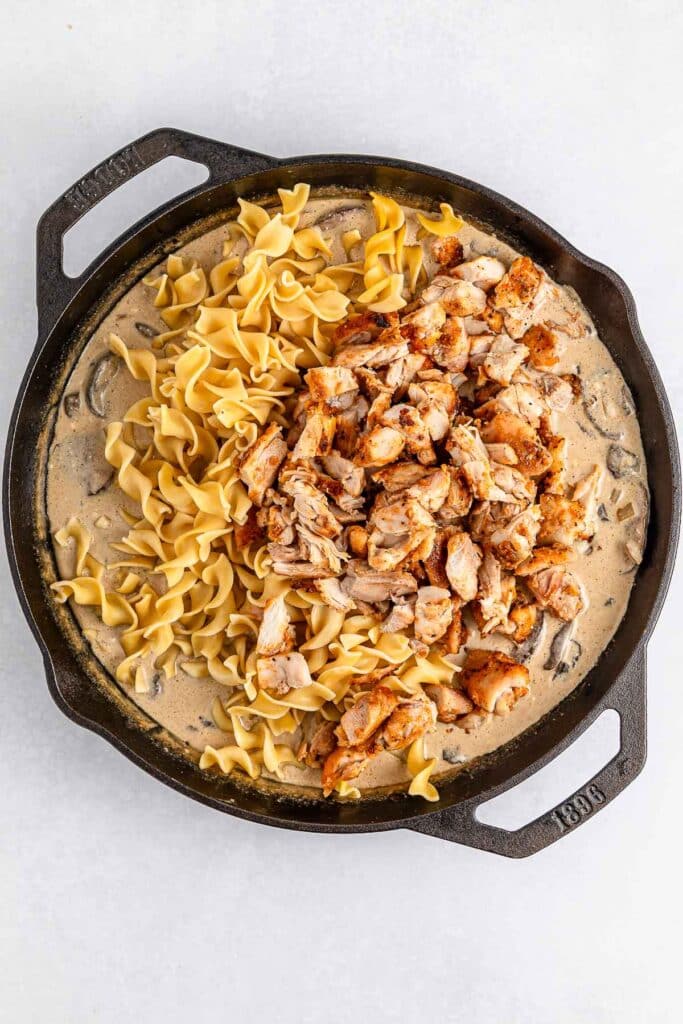 A cast iron skillet filled with creamy sauce containing mushrooms and chicken, topped with a portion of uncooked pasta.