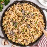 A cast iron skillet filled chicken stroganoff garnished with chopped herbs while a wooden spoon rests off to the side.