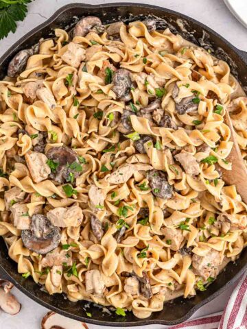 A cast iron skillet filled chicken stroganoff garnished with chopped herbs while a wooden spoon rests off to the side.