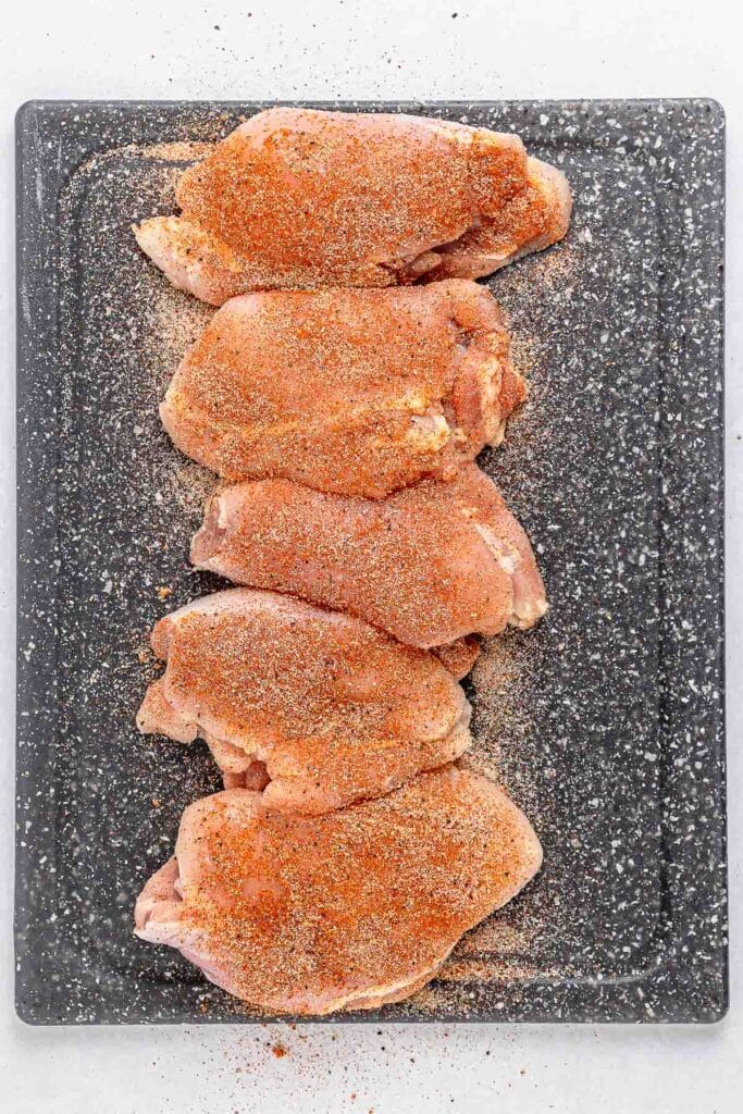 Five raw, seasoned chicken thighs arranged in a row on a cutting board.