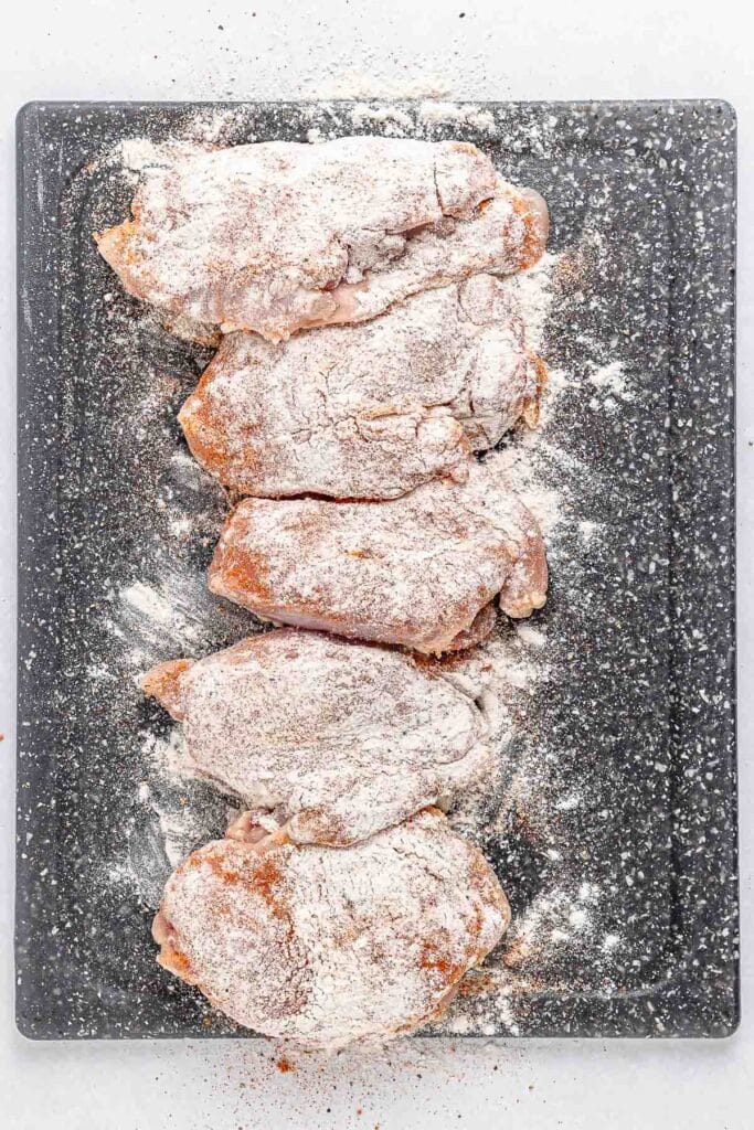 Five pieces of raw chicken thighs coated in flour are arranged on a cutting board.