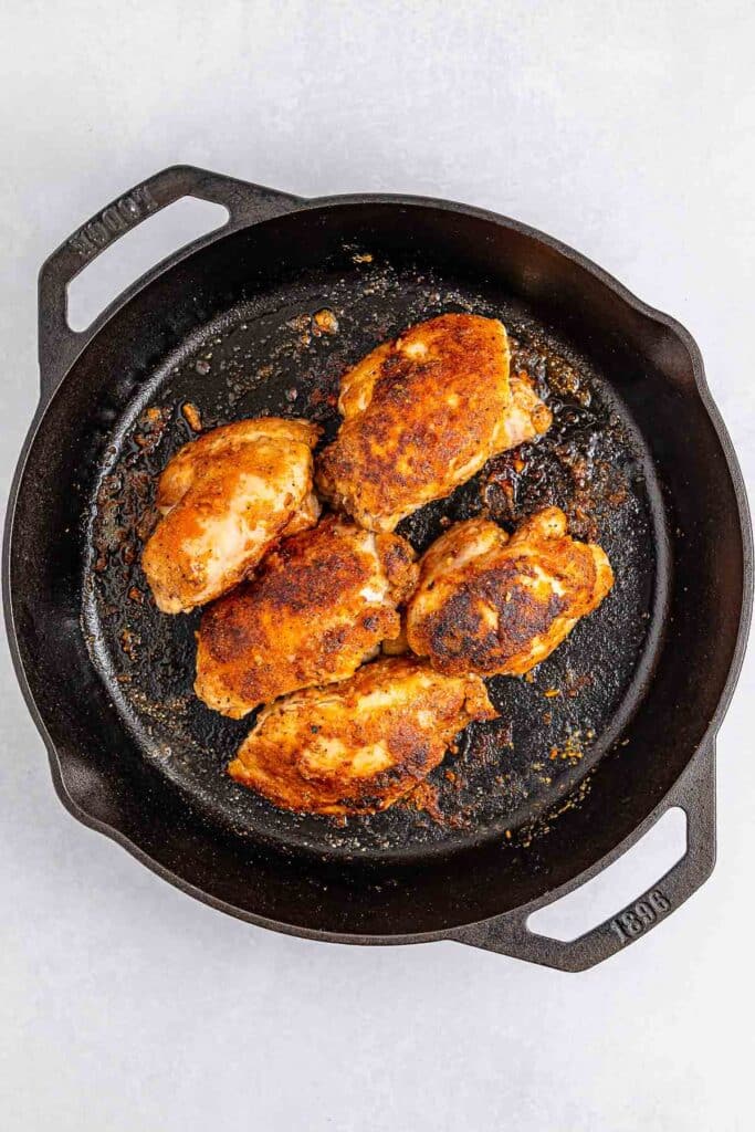 A cast-iron skillet containing five seasoned, cooked chicken thighs.