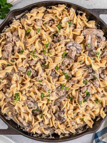 A skillet filled with creamy ground beef stroganoff garnished with chopped parsley is set on a table.