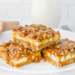 A white plate holds three pecan-topped caramel cheesecake bars. There's a glass of milk and a fork in the blurred background.