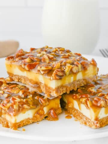 A white plate holds three pecan-topped caramel cheesecake bars. There's a glass of milk and a fork in the blurred background.