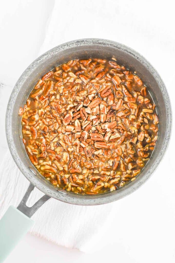 A saucepan containing chopped pecans mixed in a caramel-like sauce on a white surface.