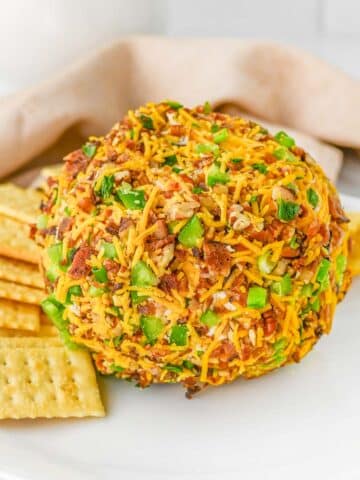 A jalapeno popper cheese ball coated with shredded cheddar cheese, chopped jalapenos and bacon bits, surrounded by rectangle crackers on a white plate.