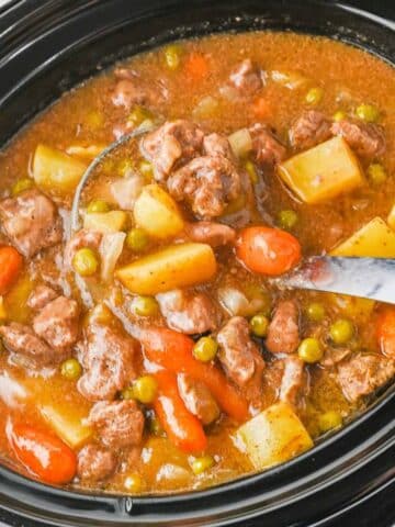 A close-up of a slow cooker filled with beef stew, featuring chunks of beef, potatoes, carrots, peas, and onions in a rich broth.