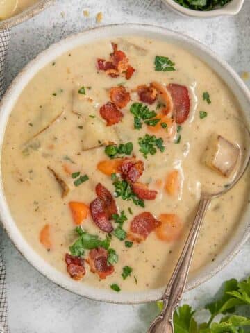 A bowl of creamy New England clam chowder garnished with chopped herbs and crispy bacon bits. A spoon is placed inside the bowl, and a checkered napkin lies next to the bowl on a light-colored surface.