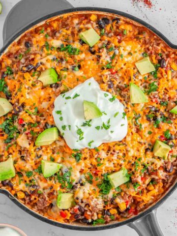 A skillet filled with mexican beef and rice topped with melted cheese, avocado, and a dollop of sour cream.