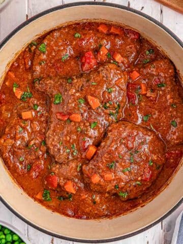 A pot of swiss steaks with carrots, tomatoes, and herbs in a red sauce.